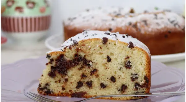 Una fetta di torta soffice con tante gocce di cioccolato, spolverata di zucchero a velo, su un piatto lilla con una forchetta.