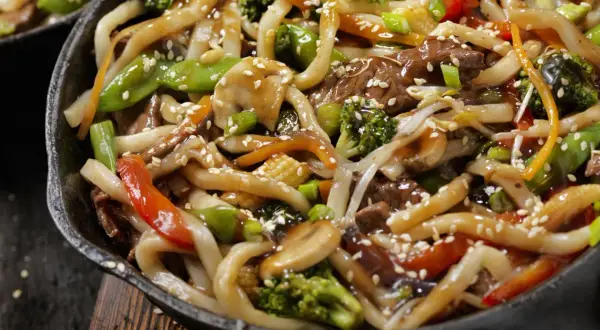Spaghetti di soia con verdure miste, carne e semi di sesamo in una padella scura.