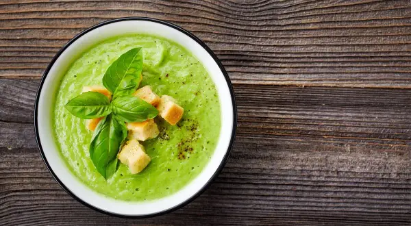 Un cremoso passato di verdure verde brillante, guarnito con crostini dorati e foglie di basilico fresco, su un tavolo di legno rustico.