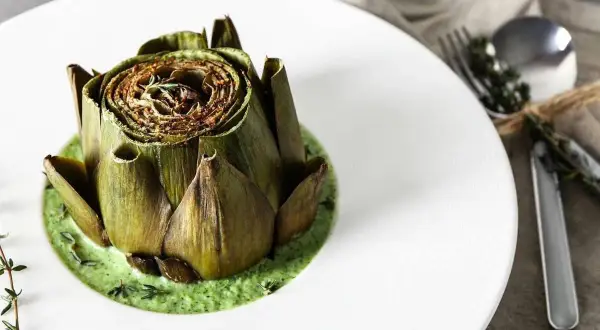 Carciofo intero cotto, adagiato su una vellutata crema di carciofi, guarnito con erbe aromatiche.