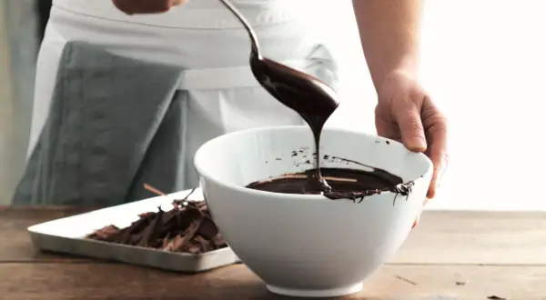 Mani che versano cioccolato fuso da un cucchiaio in una ciotola bianca, mostrando come sciogliere il cioccolato per dolci deliziosi.