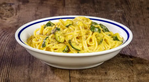 Spaghetti with zucchini and creamy sauce in a white bowl with blue trim on a wooden table.