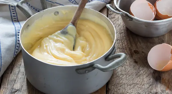Crema pasticcera veloce in pentola, con cucchiaio di legno che mescola. Guscio d'uovo accanto, pronto per un dolce.