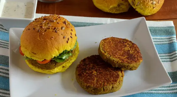 Hamburger di lenticchie dorato con semi, servito su un piatto bianco con due polpette aggiuntive e una ciotola di salsa.