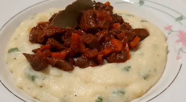 Piatto di purè cremoso con pezzetti di coratella di agnello stufata e una foglia di alloro.