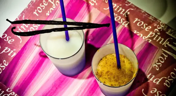 Due bicchieri con latte di riso, uno bianco e uno con spezie dorate, adornati da baccelli di vaniglia e cannucce blu su sfondo rosa.