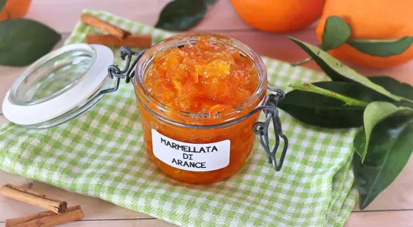 Barattolo di marmellata di arance fatto in casa, con arance fresche, foglie verdi e bastoncini di cannella sullo sfondo.