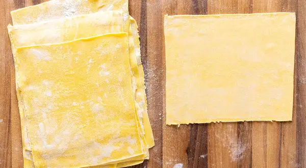 Fogli di pasta gialla, pronti per diventare deliziose lasagne fatte in casa. Un foglio singolo è posato a destra, mentre una pila di altri è a sinistra.