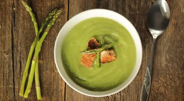 Crema di asparagi vellutata, guarnita con crostini e asparagi freschi, su un tavolo di legno rustico.