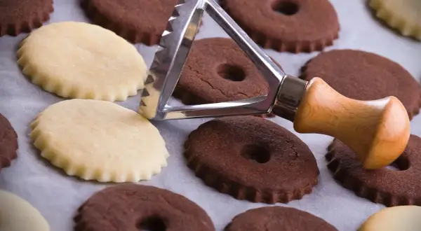 Biscotti al cacao e chiari, alcuni con foro centrale, pronti per essere infornati. Un tagliabiscotti con manico in legno è appoggiato su di essi.