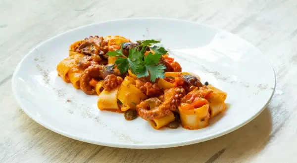 Piatto di paccheri con ragù di polpo, pomodorini e capperi, guarnito con prezzemolo fresco.