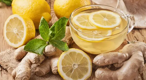 Una tazza di tisana zenzero e limone, con limoni freschi, zenzero e foglie di menta su un tavolo di legno.
