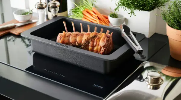 Carrè di agnello pronto per la cottura in una teglia Miele su piano a induzione. Carote e rosmarino sullo sfondo.