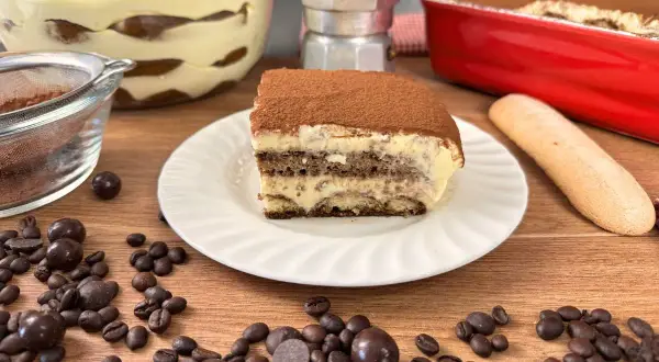 Una fetta di tiramisù con uova pastorizzate, cosparso di cacao, su un piatto bianco, circondato da chicchi di caffè e cioccolatini.