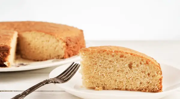 Una fetta di torta senza lievito, soffice e dorata, attende su un piatto bianco con una forchetta antica.
