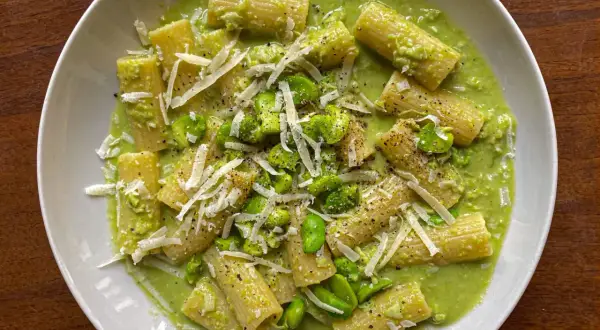Rigatoni conditi con una cremosa salsa verde e fave fresche, guarniti con scaglie di parmigiano e pepe nero. Un piatto di pasta e fave che profuma di primavera.