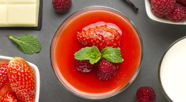 Un bicchiere di coulis di fragole, guarnito con fragola, lamponi e menta. Accanto, fragole fresche, cioccolato bianco, bacca di vaniglia e latte.