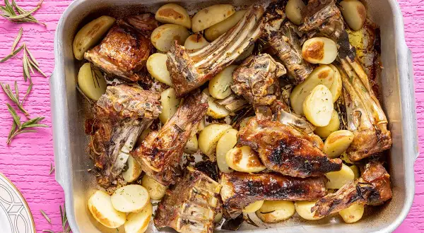 Succulento abbacchio al forno con patate dorate, profumato al rosmarino, servito in una teglia d'argento su un tavolo rosa acceso.