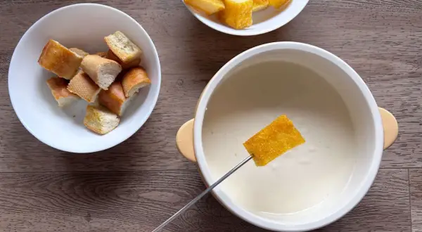 Una fetta di pane croccante viene intinta in una cremosa fonduta di parmigiano, accanto a ciotole di crostini.