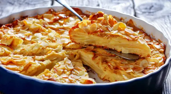 A slice of golden-brown apple cake with thin apple layers, topped with toasted almond slivers, is lifted from a blue baking dish.