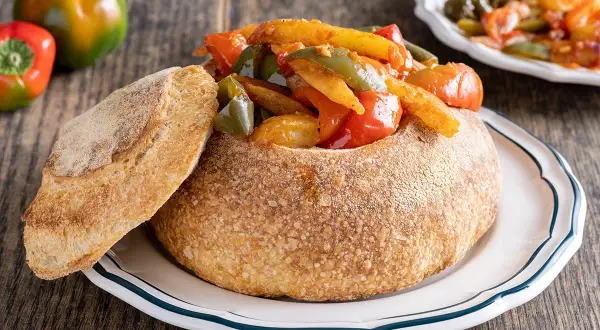 Un pane scavato ripieno di un saporito stufato di pipi e patate, con una fetta di pane croccante a lato.