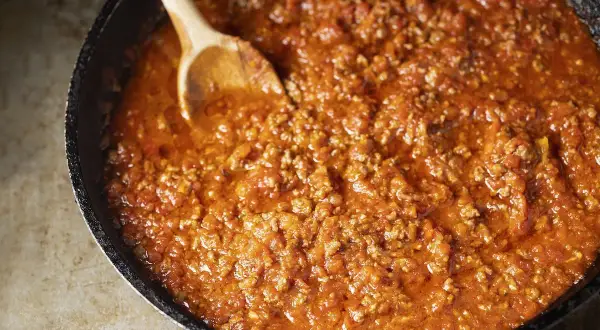 Un saporito ragù di carne in una padella, pronto per essere servito. Un cucchiaio di legno mescola la salsa rossa e densa.