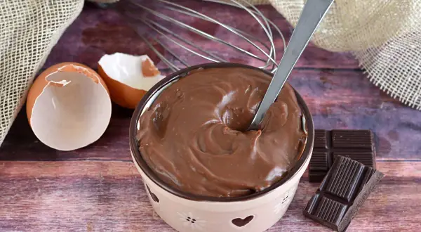 Ciotola piena di deliziosa crema al cioccolato, con gusci d'uovo, frusta e tavolette di cioccolato.