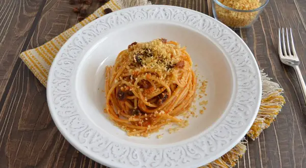 Un piatto di spaghetti con acciughe, pomodoro e pangrattato tostato, servito su un piatto bianco decorato, con un tovagliolo giallo e un bicchiere di pangrattato a lato.