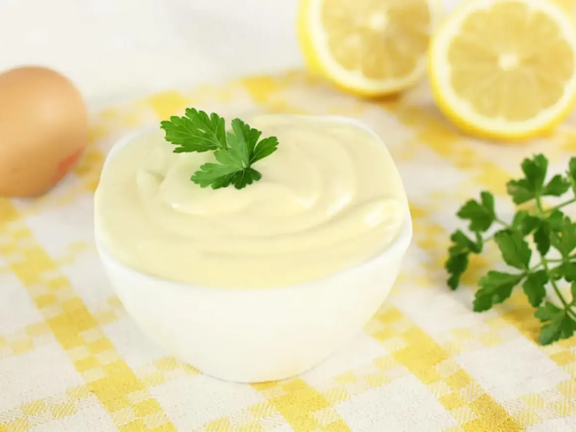 Una ciotola di cremosa maionese fatta in casa, guarnita con prezzemolo. Ingredienti freschi come uovo e limone sullo sfondo.