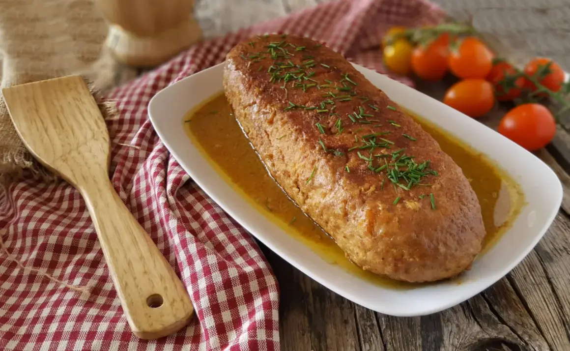 Un succulento polpettone in padella, dorato e profumato, adagiato su un letto di salsa e guarnito con erba cipollina. Accanto, una spatola in legno e pomodorini freschi.