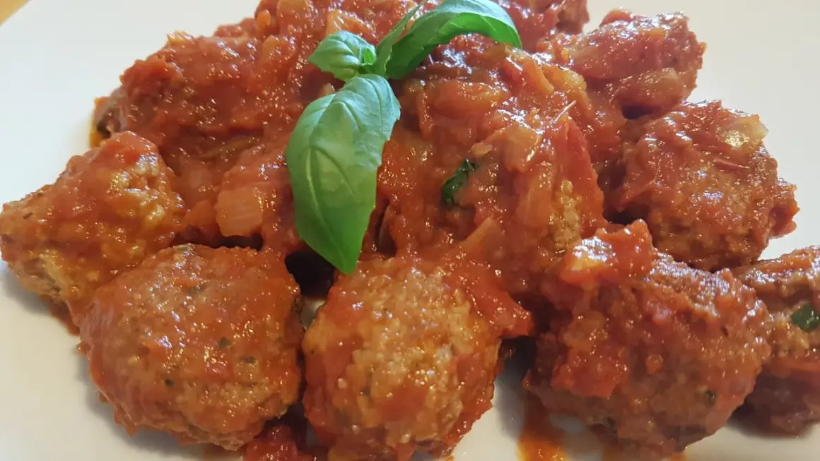 Polpette di carne morbide in un sugo rosso ricco, guarnite con foglie di basilico fresco.