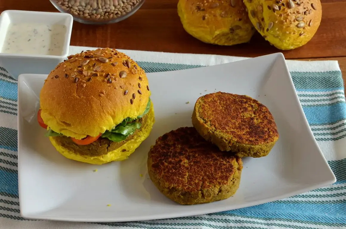 Hamburger di lenticchie dorato con semi, servito su un piatto bianco con due polpette aggiuntive e una ciotola di salsa.