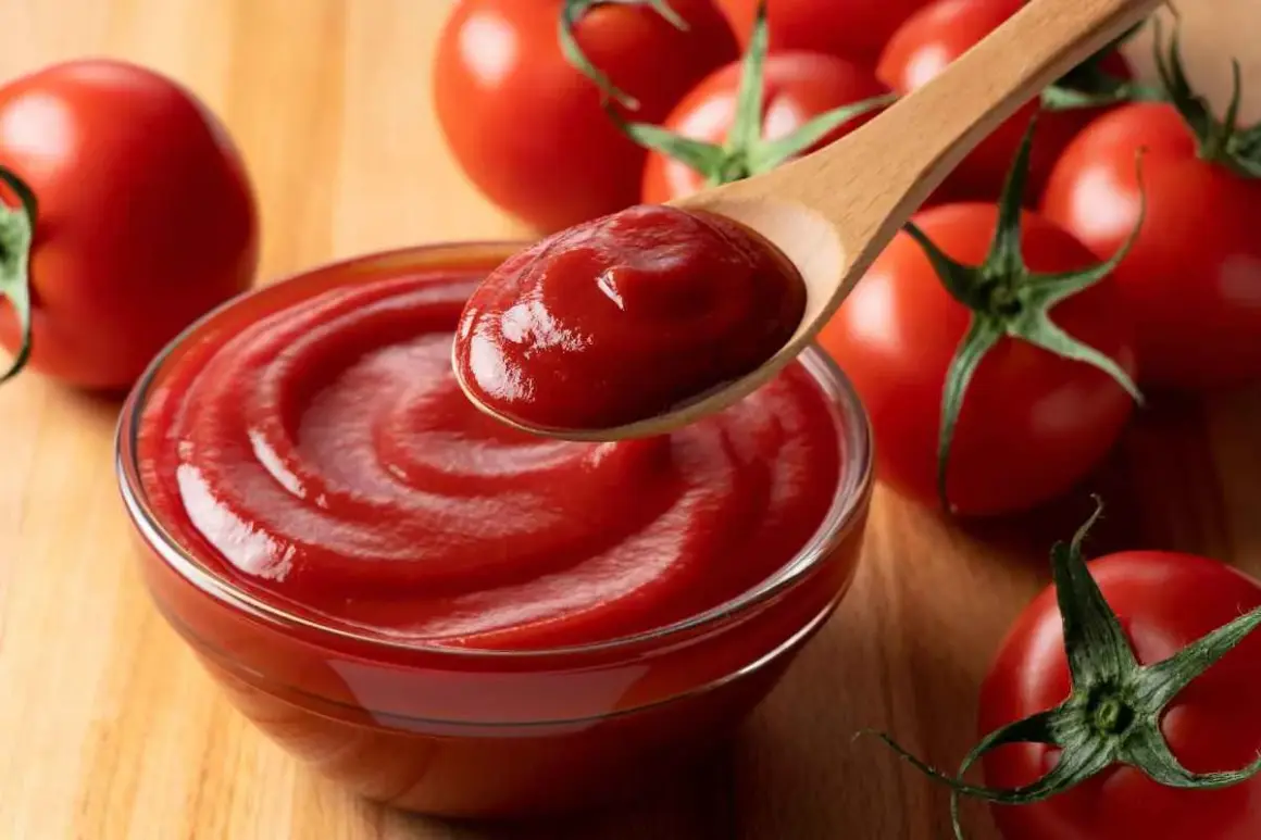 Un cucchiaio di cremoso ketchup fatto in casa, pronto per essere gustato, circondato da pomodori freschi su un tagliere di legno.