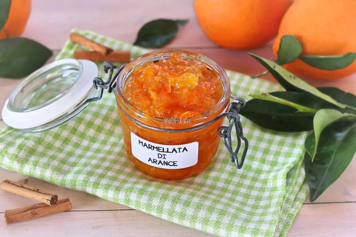 Barattolo di marmellata di arance fatto in casa, con arance fresche, foglie verdi e bastoncini di cannella sullo sfondo.