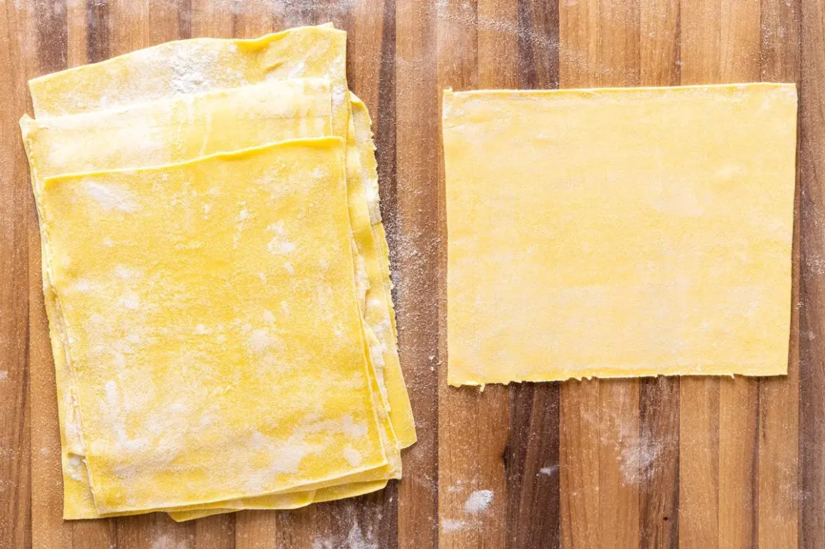 Fogli di pasta gialla, pronti per diventare deliziose lasagne fatte in casa. Un foglio singolo è posato a destra, mentre una pila di altri è a sinistra.