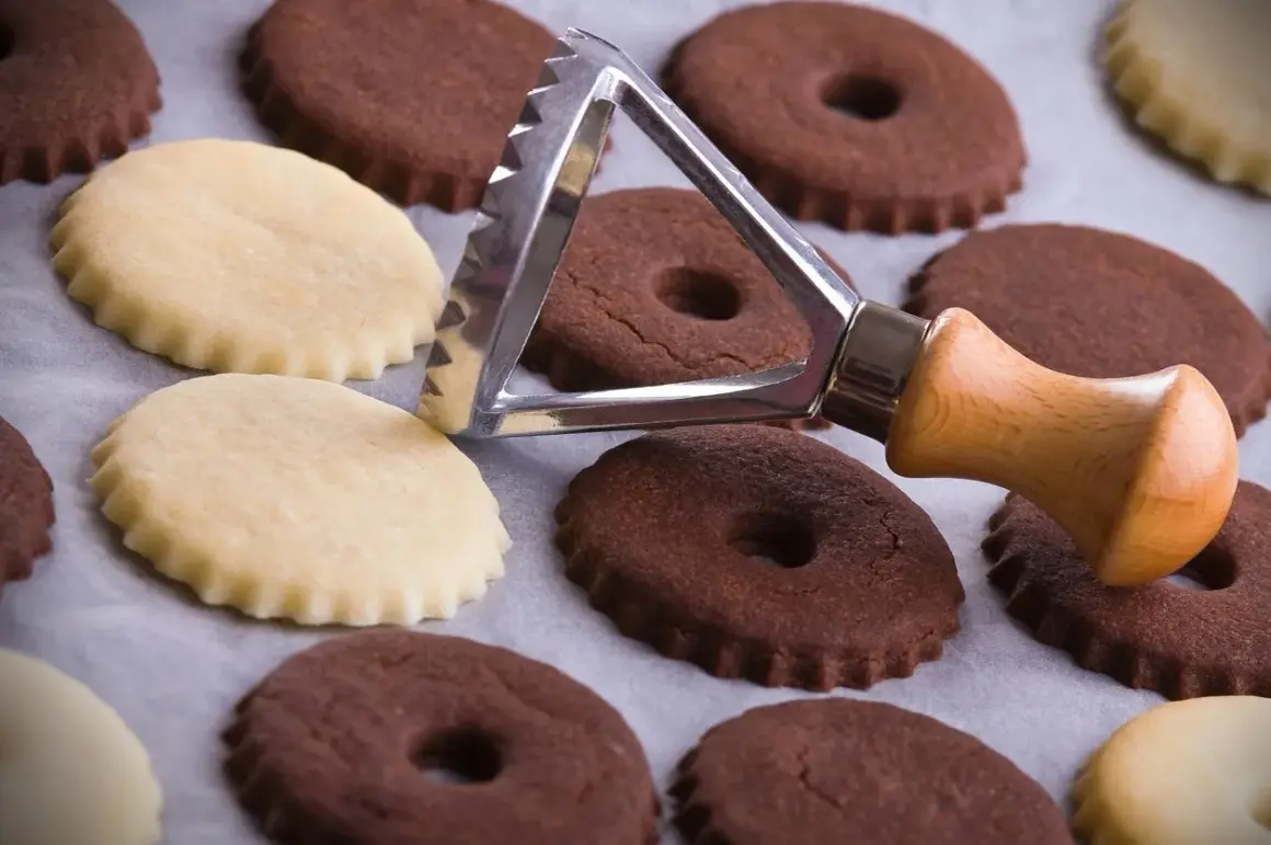 Biscotti al cacao e chiari, alcuni con foro centrale, pronti per essere infornati. Un tagliabiscotti con manico in legno è appoggiato su di essi.