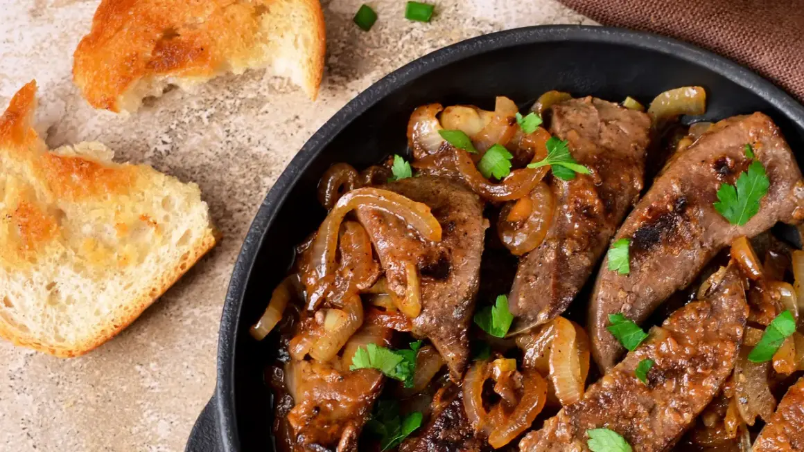 Fegato alla veneziana succulento in padella con cipolle caramellate e prezzemolo fresco, accompagnato da fette di pane tostato.
