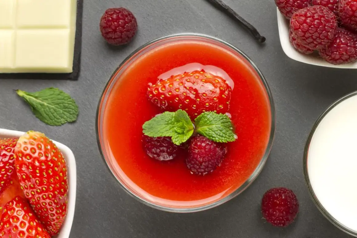 Un bicchiere di coulis di fragole, guarnito con fragola, lamponi e menta. Accanto, fragole fresche, cioccolato bianco, bacca di vaniglia e latte.