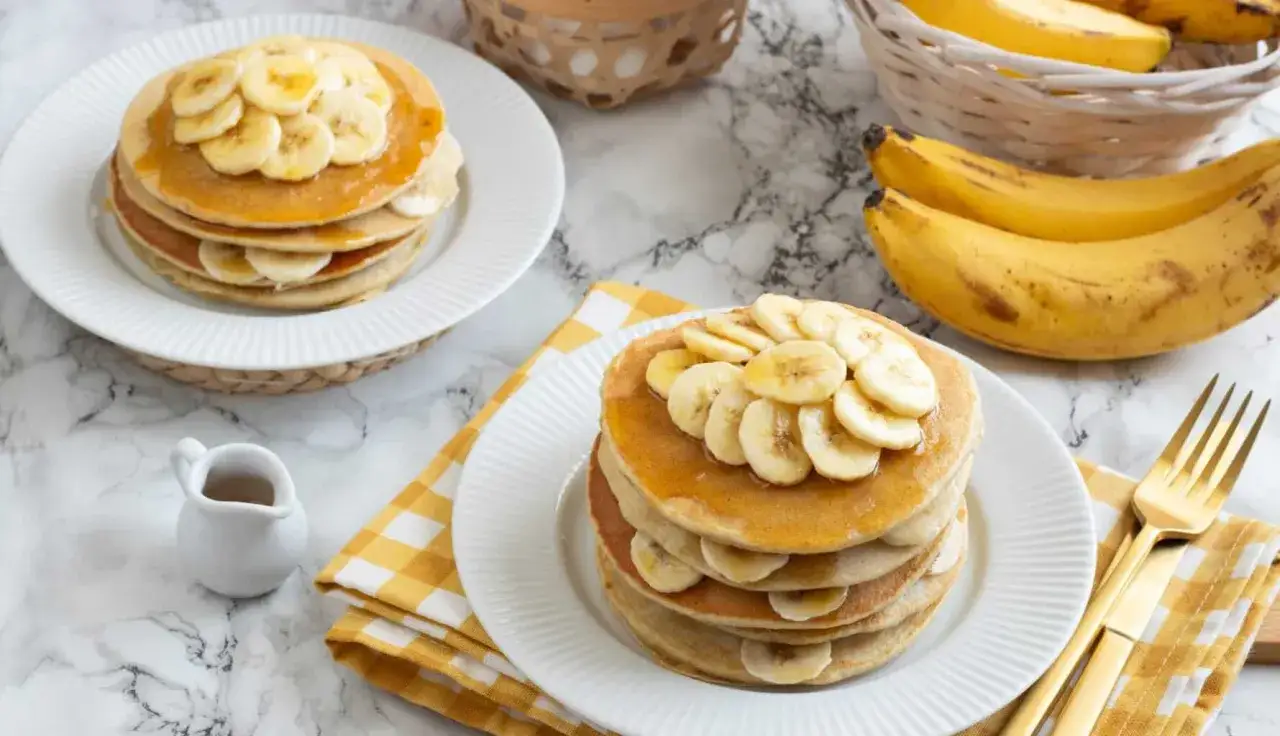 Pila di pancake alla banana, guarniti con fette di banana fresca e sciroppo dorato. Sullo sfondo, banane mature in un cestino.