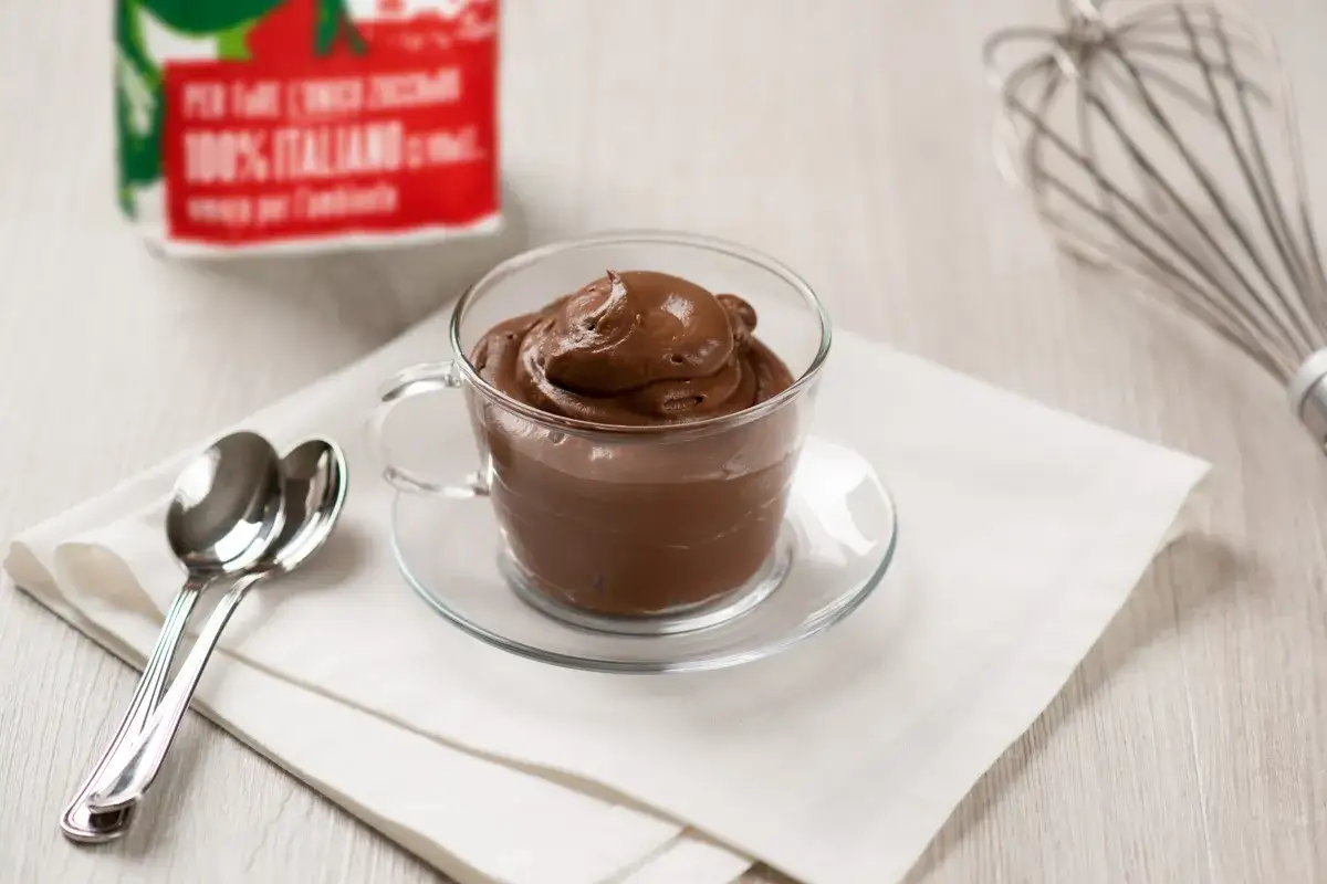 Coppa di crema pasticcera al cioccolato, pronta per essere gustata. Accanto, due cucchiaini e una frusta.