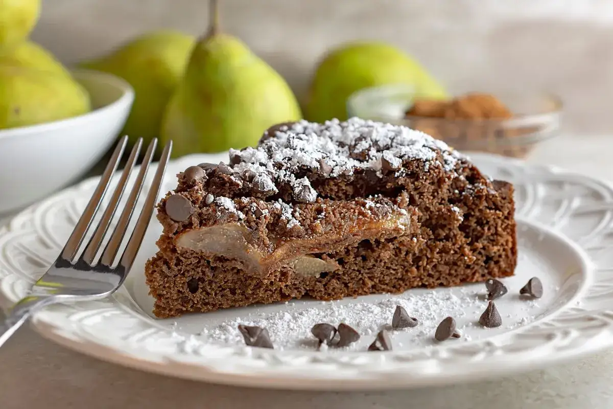 Una fetta di torta pere e cioccolato, cosparsa di zucchero a velo e gocce di cioccolato, su un piatto bianco con una forchetta.