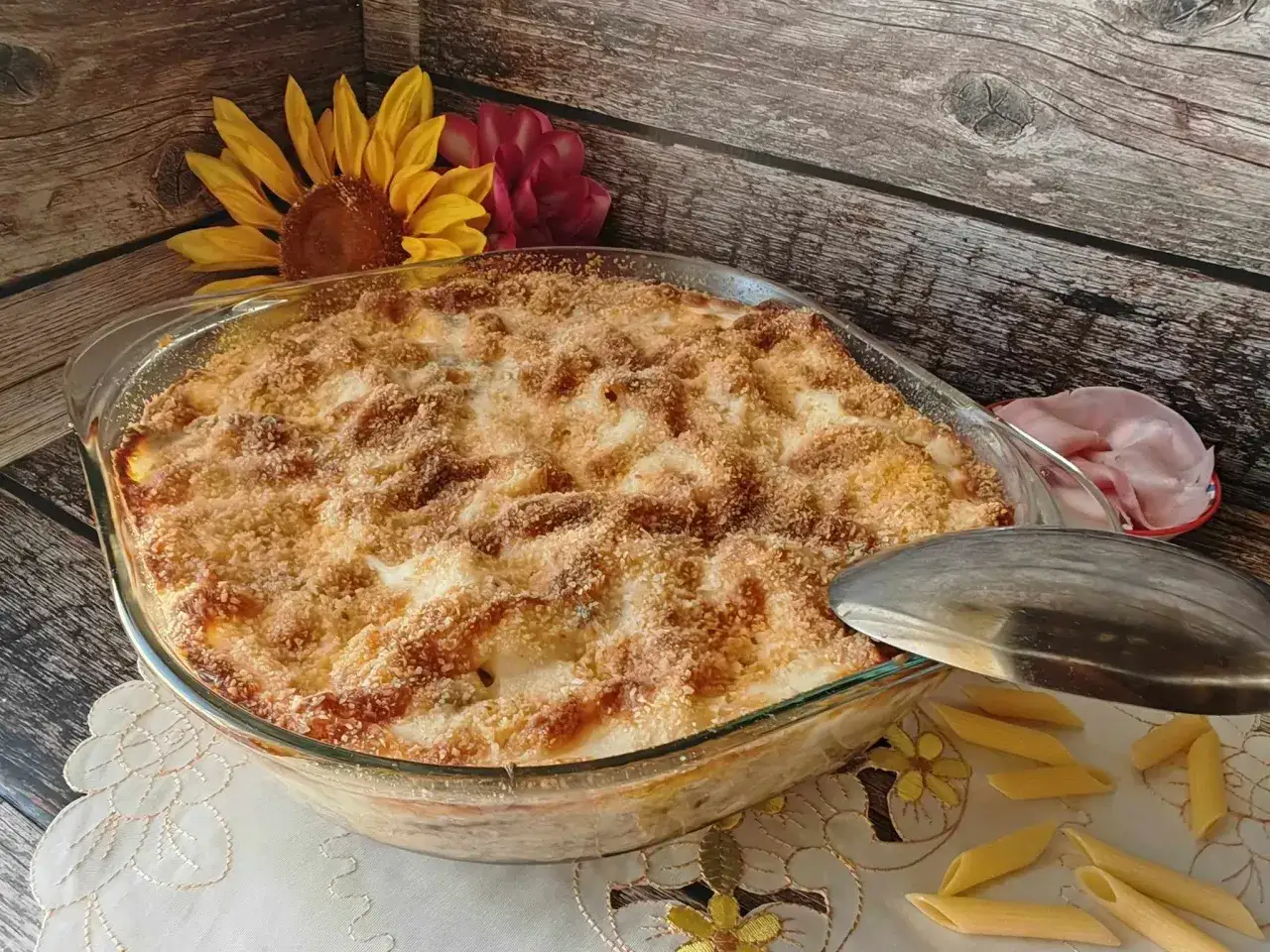 Un invitante pasta al forno dorata e croccante, pronta per essere servita. Accanto, fiori e penne crude anticipano il sapore.