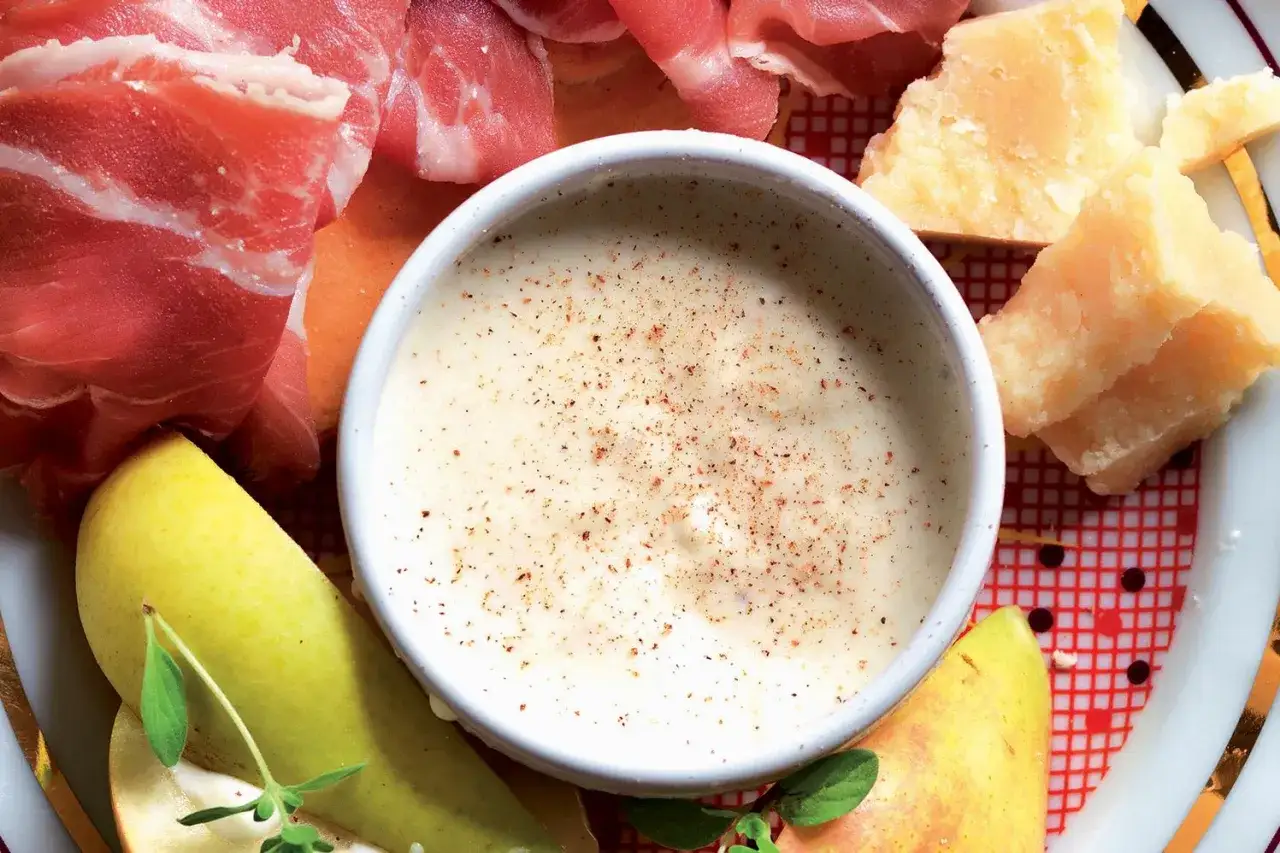 Una ciotola di cremosa fonduta di parmigiano, cosparsa di pepe, circondata da prosciutto crudo, pere e scaglie di formaggio.