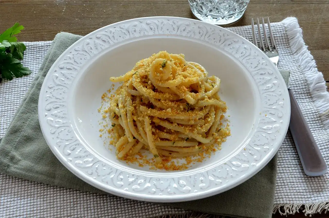 Un piatto di pasta con acciughe, condita con pangrattato dorato e prezzemolo fresco, servita su un elegante piatto bianco.