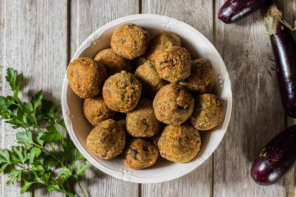 Un piatto colmo di polpette di melanzane al forno, dorate e croccanti, accanto a melanzane fresche e prezzemolo.