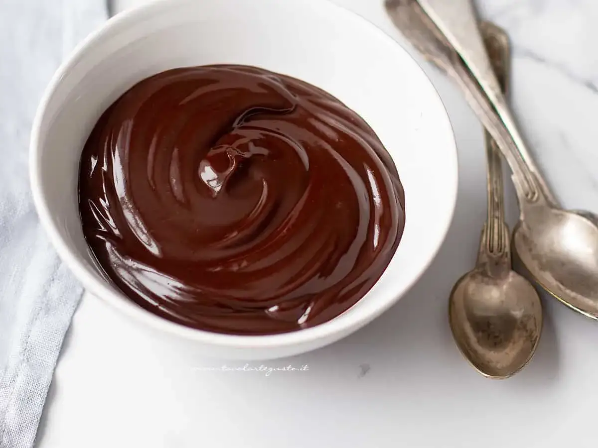 Una ciotola bianca piena di lucida glassa al cioccolato, pronta per essere gustata. Due cucchiai d'argento sono appoggiati accanto.