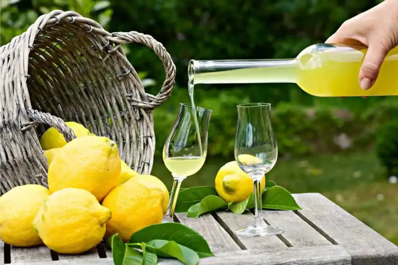 Versando limoncello ricetta originale Amalfi da una bottiglia in un bicchiere, con limoni freschi e un cesto sullo sfondo.
