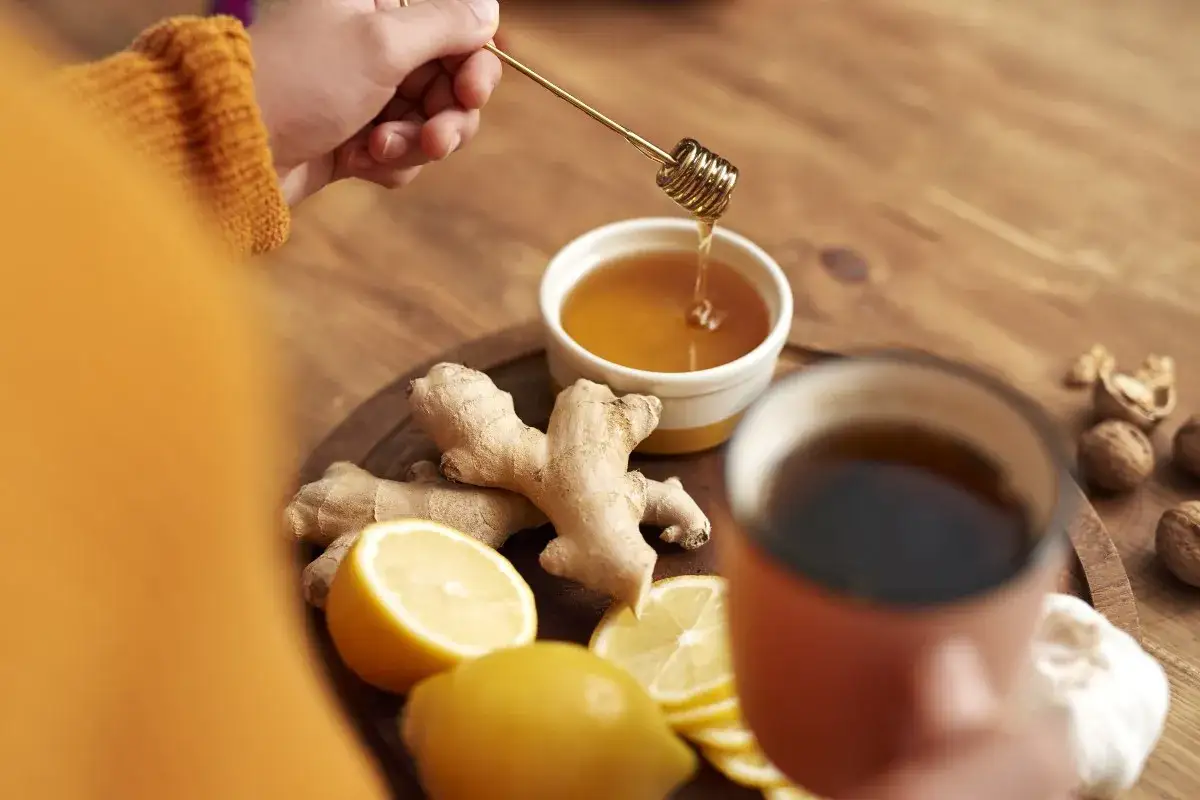 Mano aggiunge miele a una tazza di tisana zenzero e limone, con zenzero fresco, limoni e noci sul tavolo.