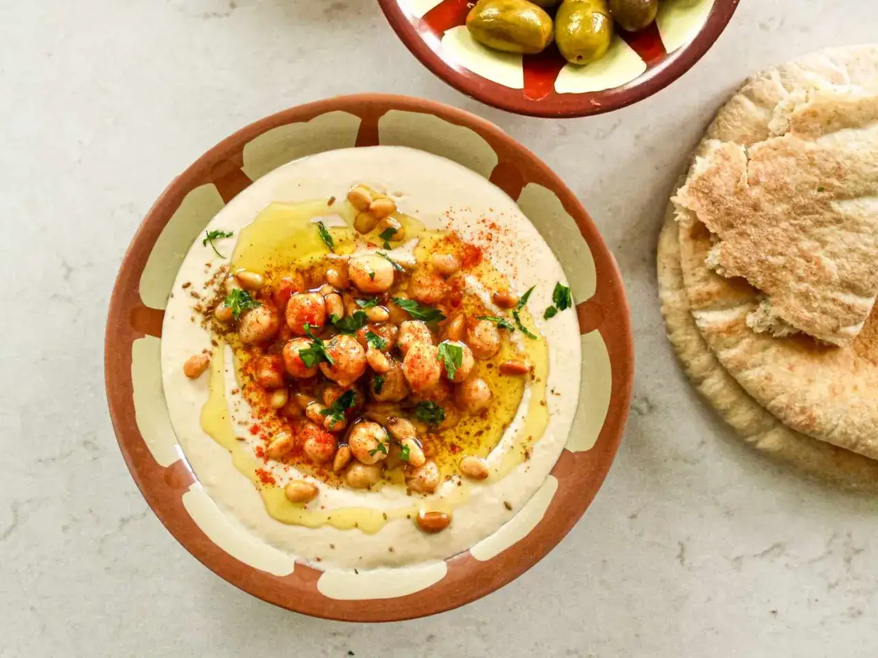 Hummus di ceci cremoso, condito con olio, paprika, pinoli e prezzemolo, servito con pane pita e olive.