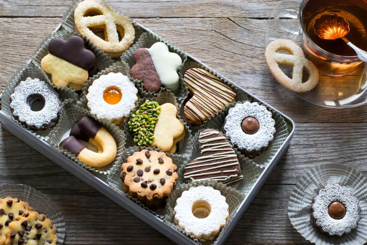 Scatola piena di biscotti pasta frolla decorati: a forma di cuore, con cioccolato, marmellata e pistacchi. Accanto, una tazza di t&egrave; caldo.
